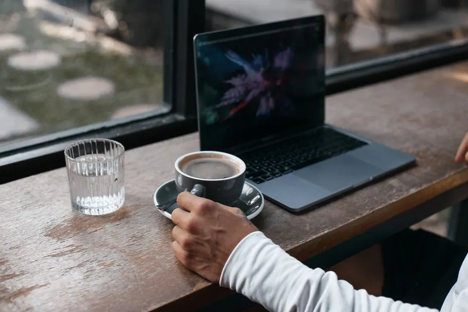 MacBook đặt cạnh tách cà phê và ly nước trên bàn gần cửa sổ