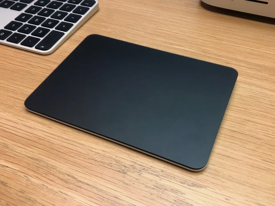 Magic Trackpad mặt màu đen đặt cạnh bàn phím trên bàn gỗ